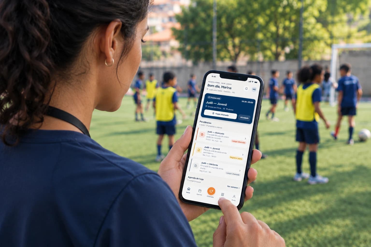 Educadora segurando o celular com o app do Cais aberto, enquanto crianças de colete jogam futebol no campo ao fundo
