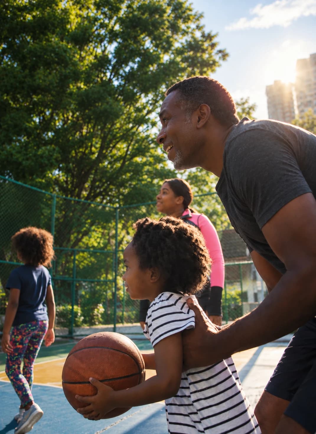 Educador agachado ao lado de uma criança que segura uma bola de basquete numa quadra ao ar livre, com outras crianças ao fundo
